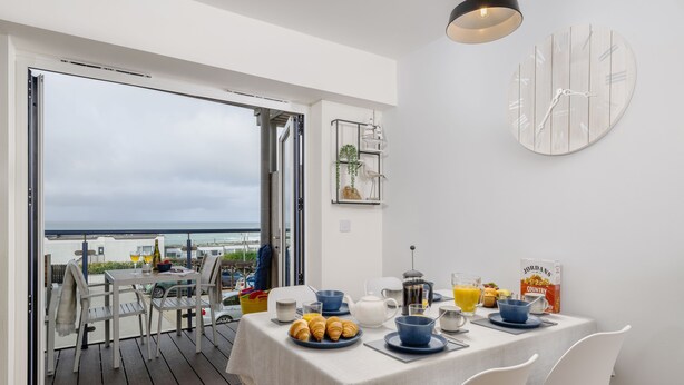 Un coin repas avec une table mise pour le petit-déjeuner, une vue sur la mer et une horloge moderne accrochée au mur.