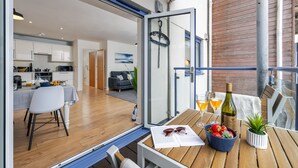 Une cuisine moderne avec des meubles blancs, un coin repas avec une table et des chaises en bois, et un balcon offrant une vue sur la ville.