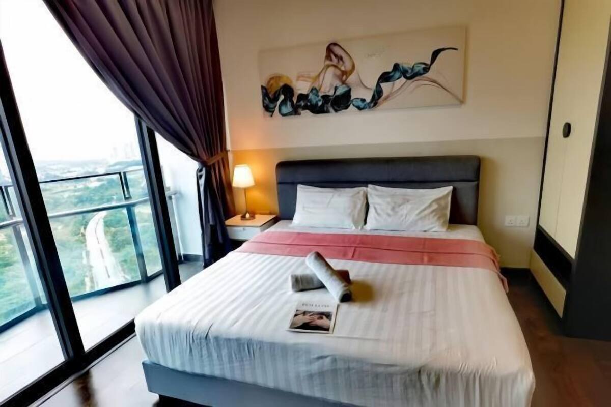 A hotel room with a large bed, a nightstand with a lamp, a painting on the wall, and a view of the city through large windows.