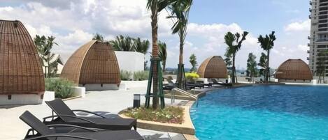 Zona junto a la piscina con sillones de descanso, palmeras y estructuras de techo de paja.