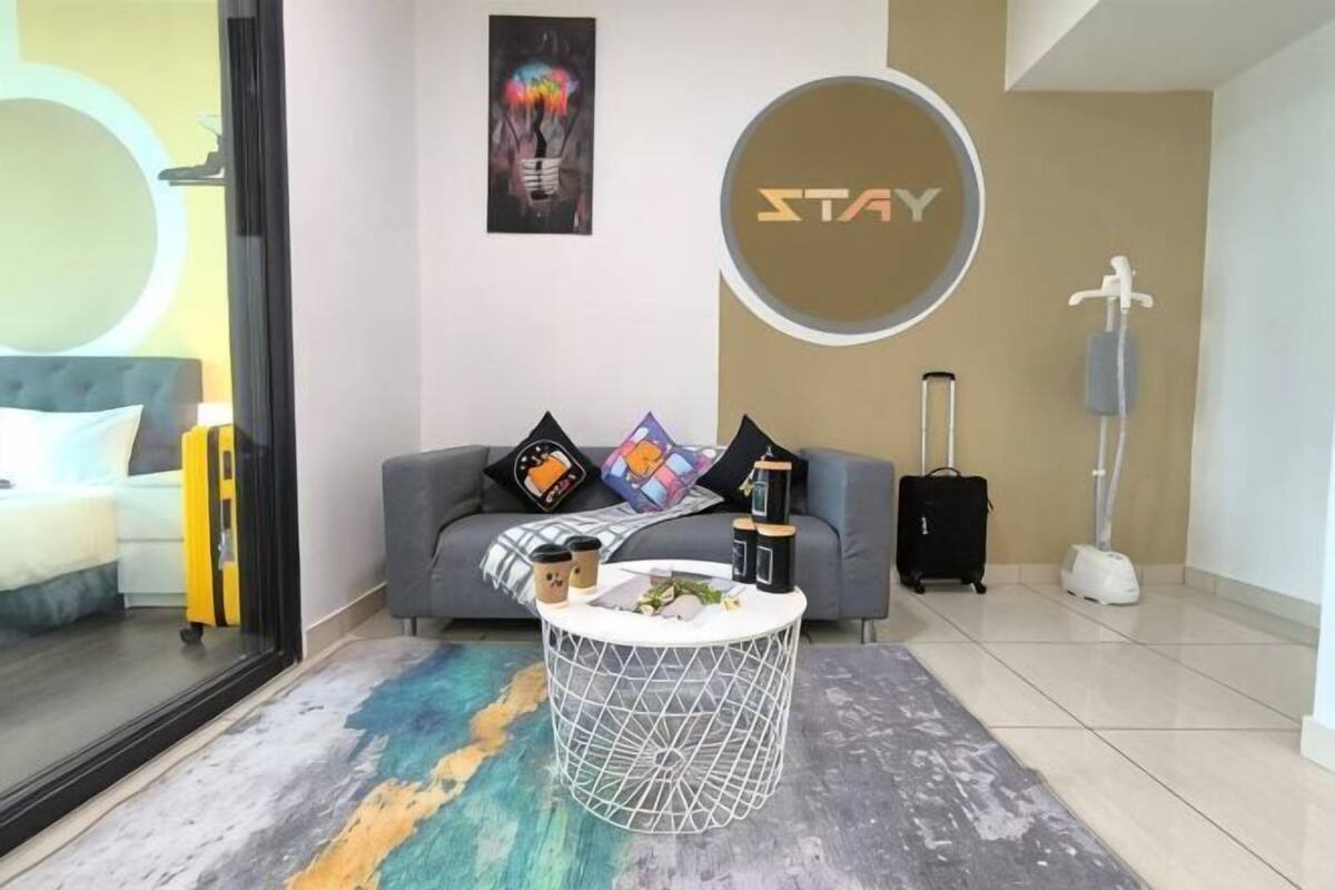 A modern hotel room with a grey sofa, a round coffee table, a large mirror, and a 'STAY' sign on the wall.