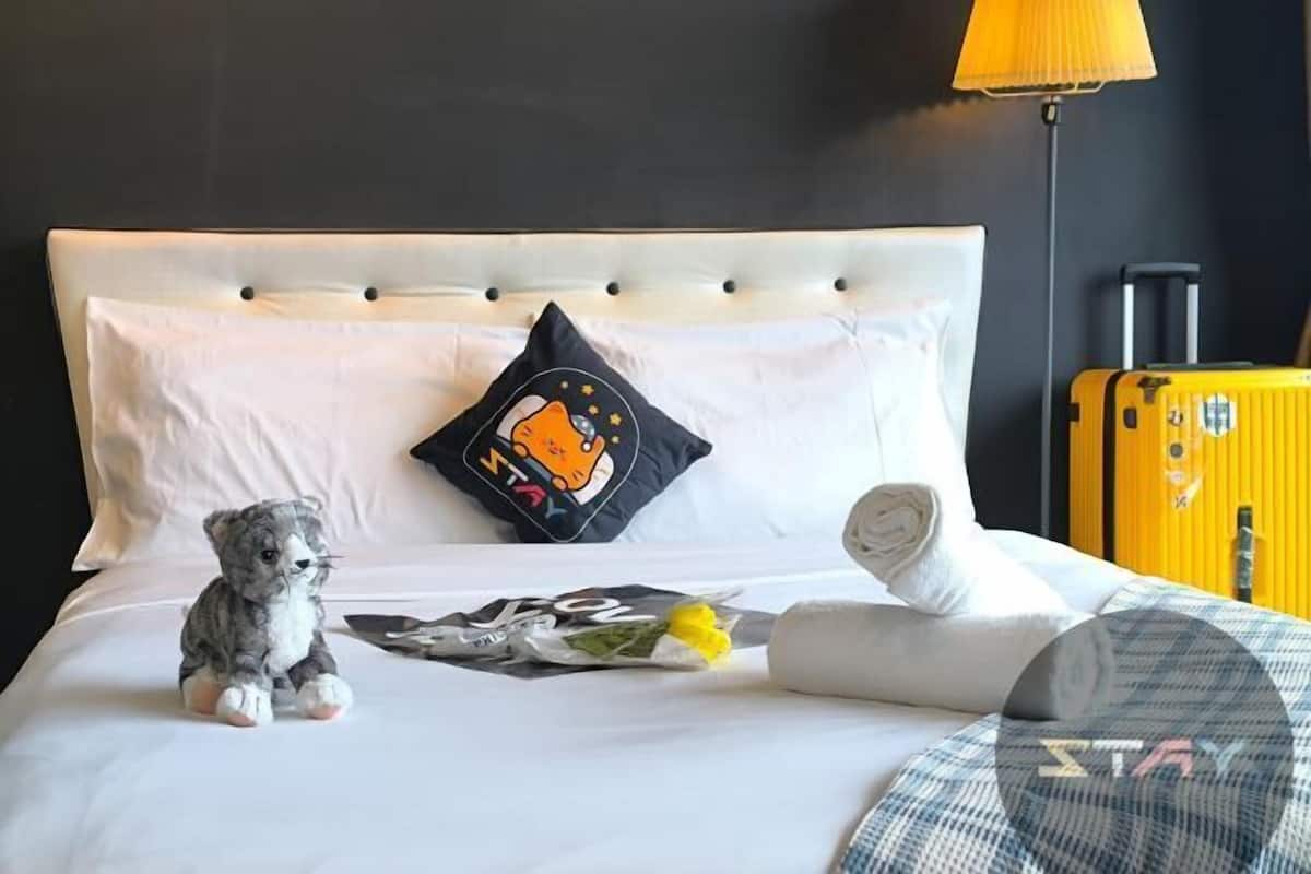A bed with a plush toy, a pillow with a tiger design, a rolled towel, and a suitcase against a dark wall.