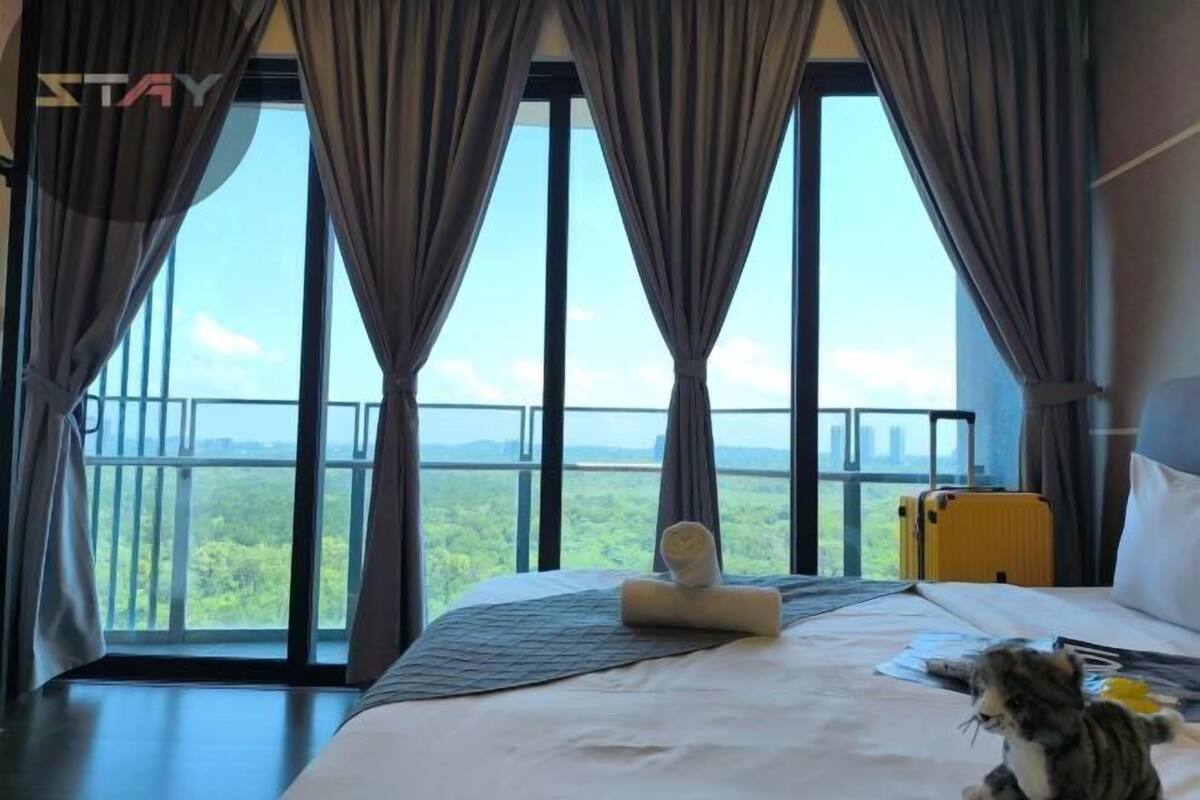 A hotel room with a large bed, a view of greenery through the windows, and a suitcase on the floor.