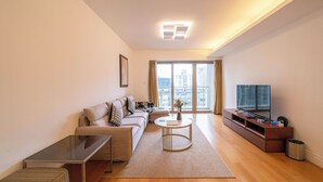 Una sala de estar moderna con un conjunto de sofás, una mesa de centro, un televisor sobre un mueble de madera y un amplio ventanal con vista a la ciudad.