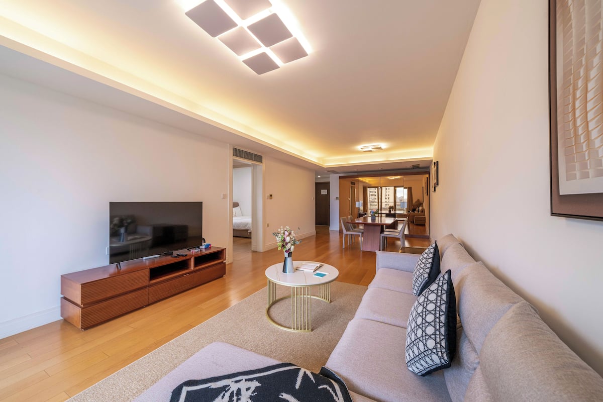 Ein modernes Wohnzimmer mit Flachbildfernseher, Holzunterhaltungseinheit und beiger Couch mit gemusterten Kissen.