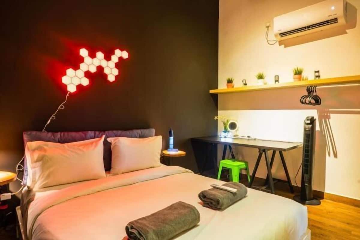 Habitación de hotel con cama, mesita de noche, escritorio, silla, unidad de aire acondicionado y estante en la pared con plantas.