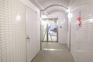 A corridor with white tiled walls and a tiled floor, leading to a door with a metal handle. - City la Defense (Sapporo)