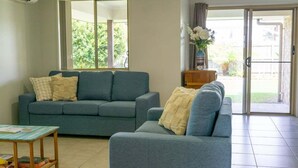 Un salón con un sofá gris, una pequeña mesa de madera y vistas a un espacio exterior a través de un ventanal corredero.