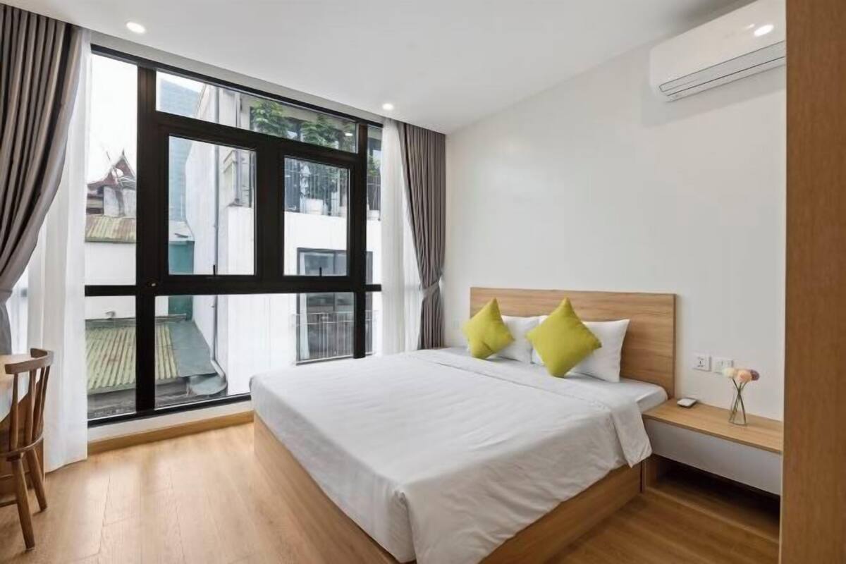 Habitación de hotel con un ventanal grande, una cama con ropa blanca y almohadas verdes, una mesita de noche de madera y una pequeña mesa con una silla.