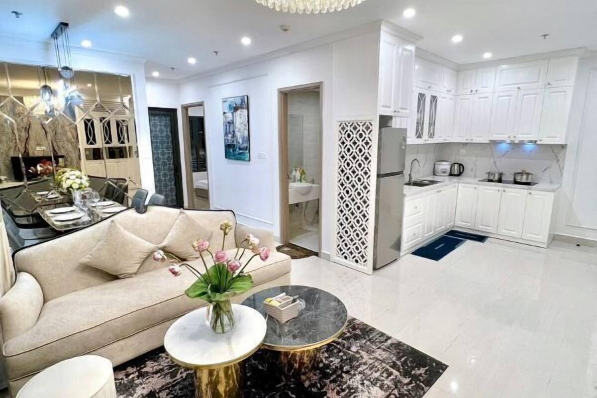 Sala de estar moderna com sofá, mesa de centro e um vaso decorativo. Ao lado, cozinha com armários brancos e um espaço de jantar.