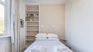 A neatly arranged bedroom with a bed, pillows, a bedside table, a shelf with books and a plant, and a window with curtains. - Stylish West London Stay by Hammersmith (London)