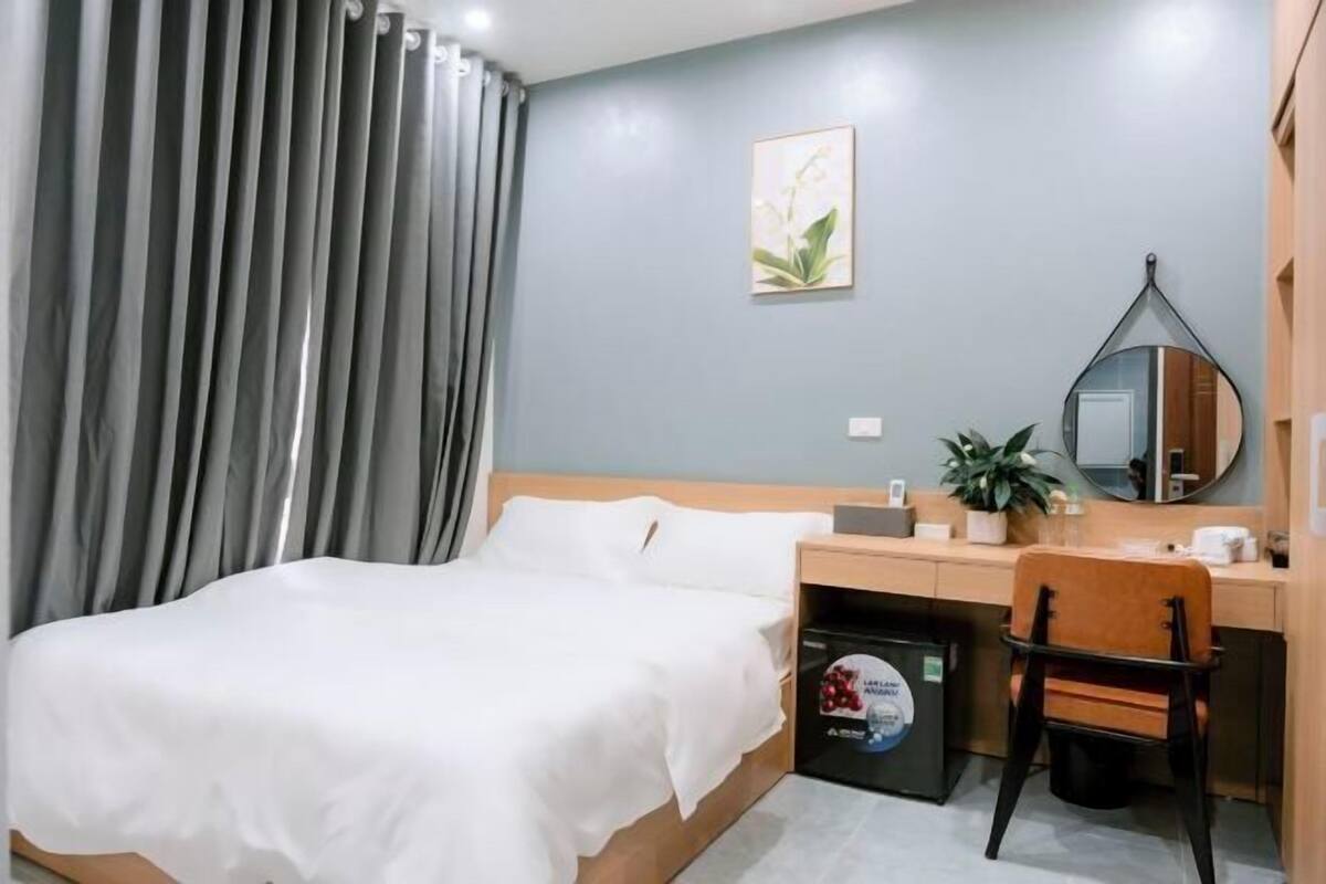 Une chambre d’hôtel comprenant un lit, un bureau avec une chaise, un miroir, une plante en pot et un petit réfrigérateur.