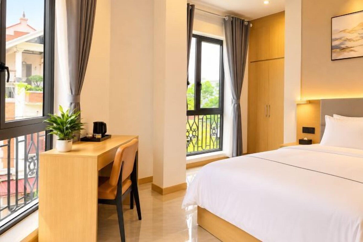 A hotel room with a bed, a desk with a chair, a potted plant, and a view of the outside.