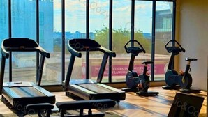 Une salle de sport équipée de tapis de course, de vélos elliptiques et d’un espace avec des bancs, avec de grandes fenêtres offrant une vue sur les bâtiments et les arbres.