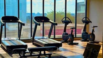 A gym with treadmills, elliptical machines, and a stationary bike, with a view of a cityscape through large windows.