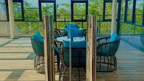 Un espace extérieur aménagé avec des tables rondes et des chaises en osier, protégé par un auvent et entouré de panneaux de verre, offrant une vue sur la verdure.