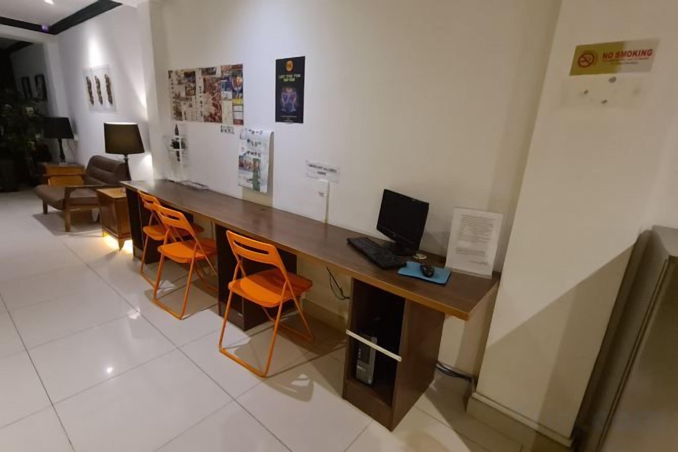 Quarto com uma escrivaninha de madeira, um laptop e três cadeiras laranjas. Há decorações nas paredes e um aviso de 'No Smoking'.