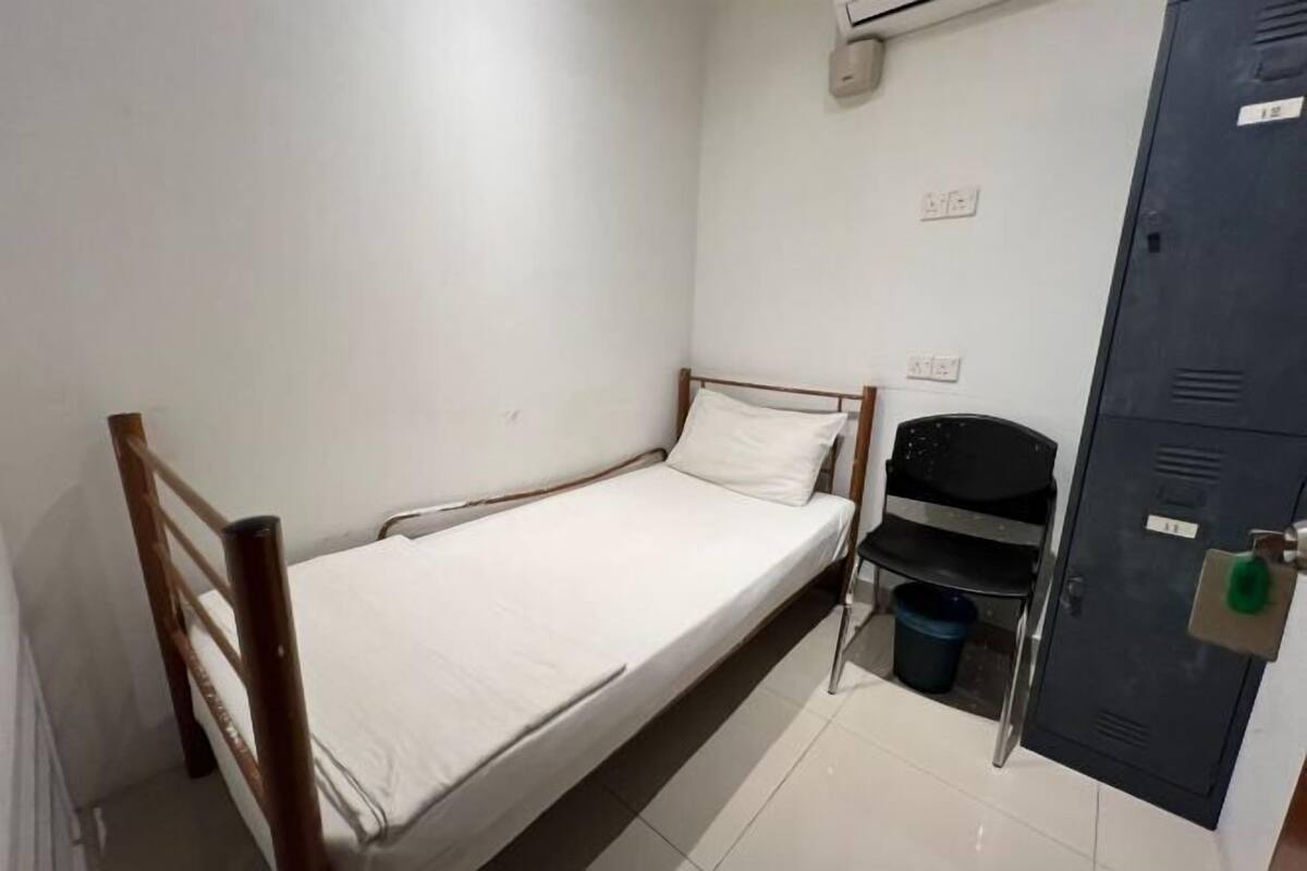 Une petite chambre avec un lit simple, un cadre en bois, un matelas blanc, une chaise noire, un seau bleu et un casier en métal.