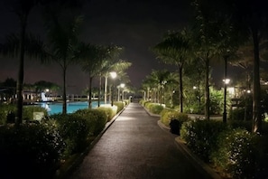 Un chemin menant à une zone de piscine la nuit, bordé de palmiers et éclairé par des lampadaires.