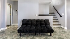 A black sofa in a room with patterned floor tiles, a staircase, and a door. - Modern 3BR Bay Stay: pet/family-friendly, yard, parking, W/D, near hospitals (MORENO VALLEY)