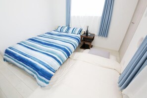 A single bed with a striped blue and white bedspread, a bedside table with a lamp, and light blue curtains. - Nishi-Shinjuku-Villa (Tokyo)