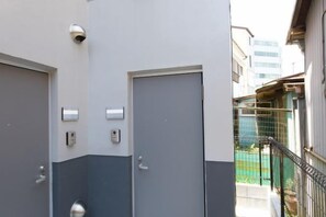 A building exterior with two doors, a security camera, and a fenced area with a building in the background. - Nishi-Shinjuku-Villa (Tokyo)