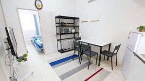 A compact room with a bed, a kitchenette, a dining area with a table and chairs, a TV, and a shelf with kitchen items. - Nishi-Shinjuku-Villa (Tokyo)