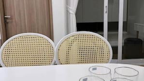 Une pièce avec une table blanche, deux chaises en osier et une porte vitrée donnant sur un balcon.