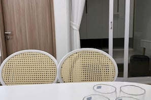 Une pièce avec une table blanche, deux chaises en osier et une porte vitrée donnant sur un balcon.