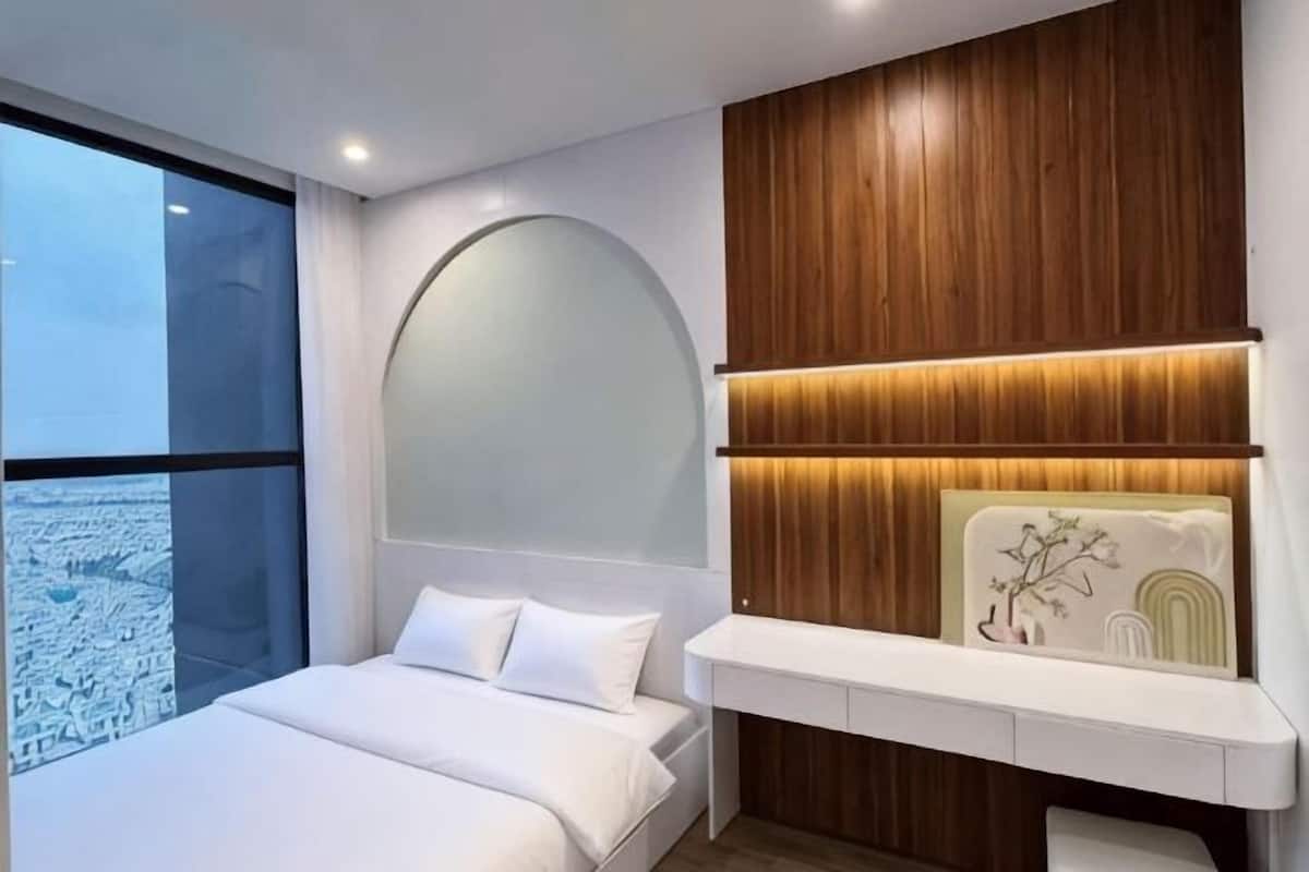 A modern bedroom with a large bed, a wooden panel wall, a built-in shelf, and a large window with a city view.