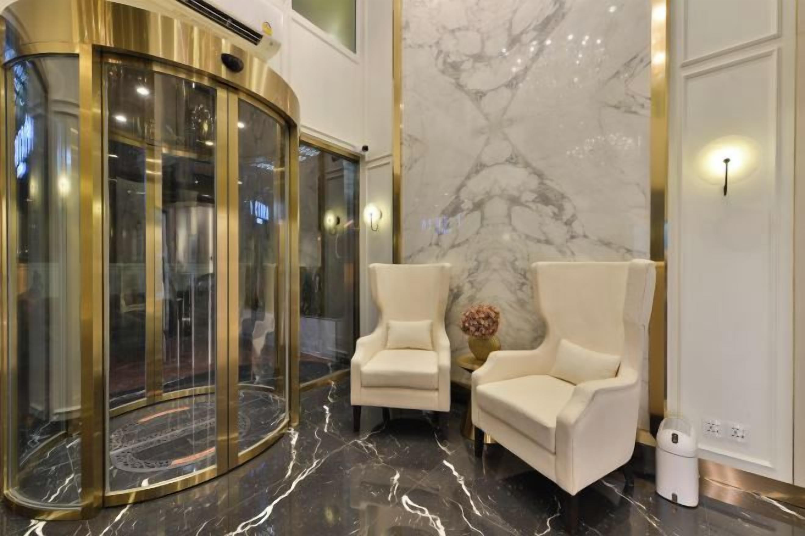A lobby with a marble wall, two white armchairs, a gold elevator, and a marble floor.