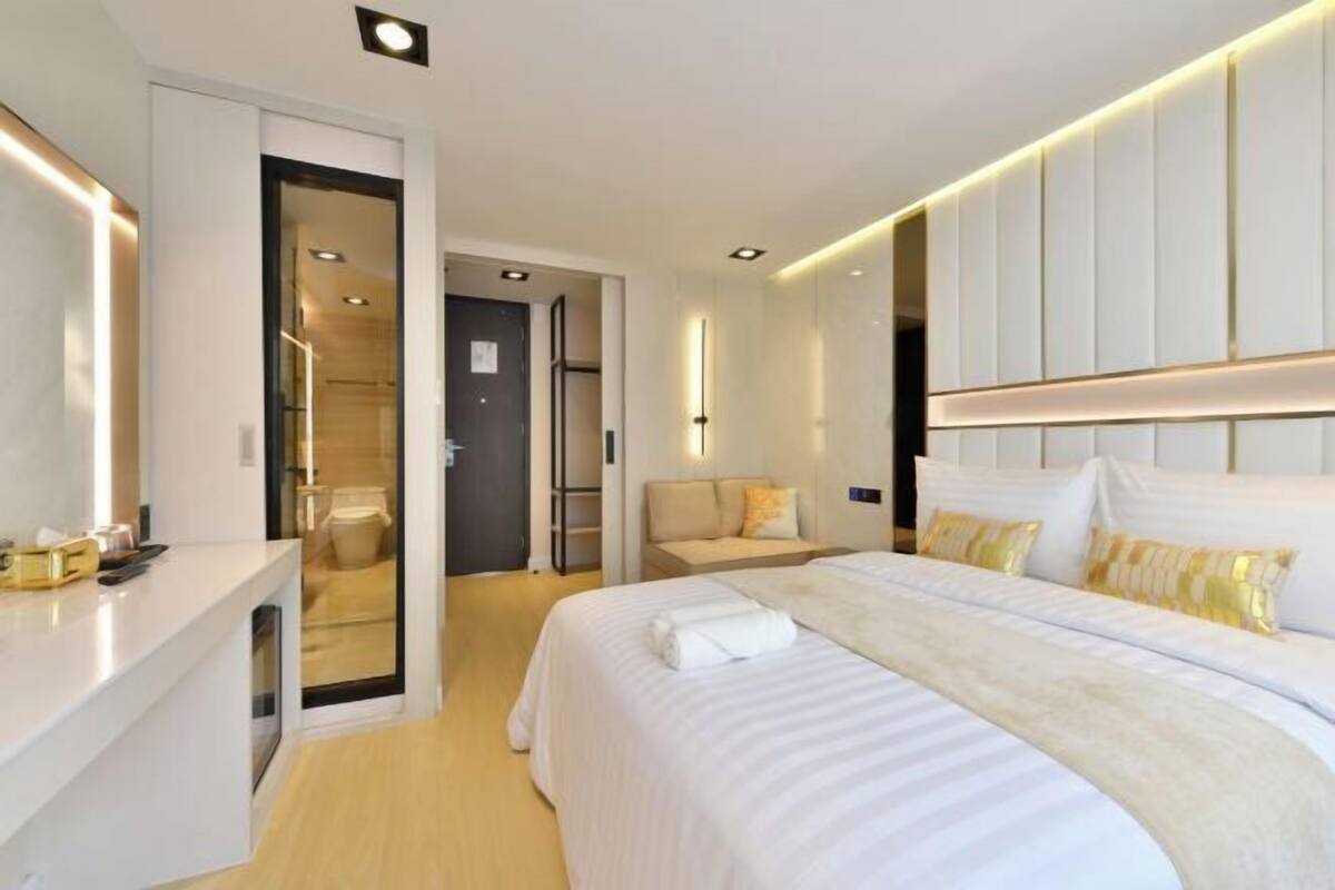 A modern hotel room with a large bed, a bedside table, a walk-in closet, and a bathroom visible through an open door.