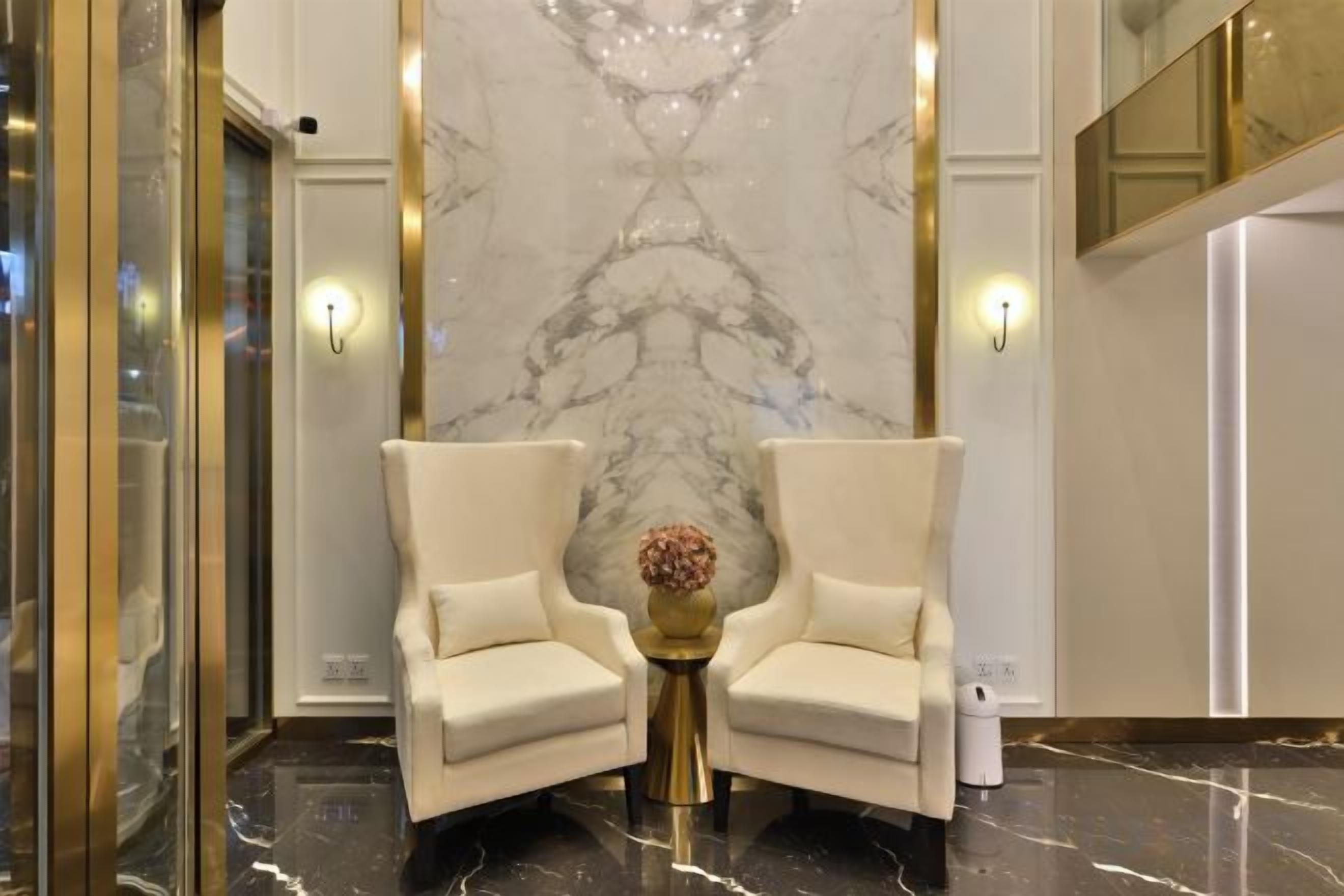 A lobby with two white armchairs, a marble wall, and a small table with a vase.