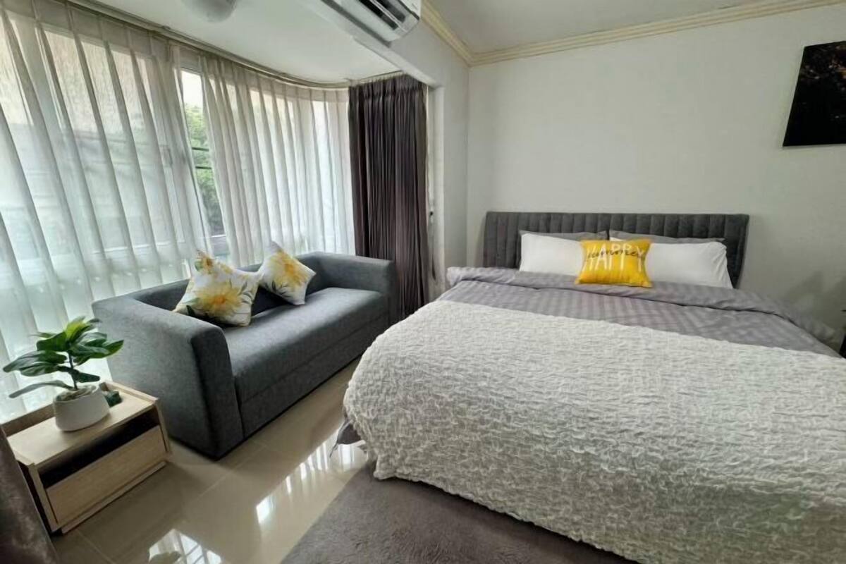 A hotel room with a bed, a grey sofa, a small table with a plant, and a window with curtains.