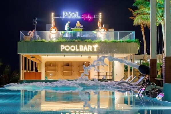 Un bar junto a la piscina con un letrero de neón, una piscina con juegos de agua y la fachada de un edificio moderno.
