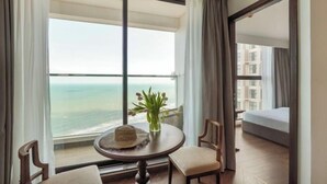 A hotel room with a large window offering a view of the ocean, a round table with a hat and flowers, and a bed. - Mandala Heaven Dreamscape Hotel & Resort (Phan Thiet)