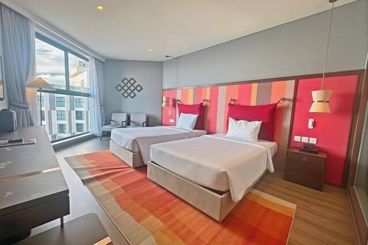 A hotel room with two beds, a desk, a chair, a TV, and a large window.