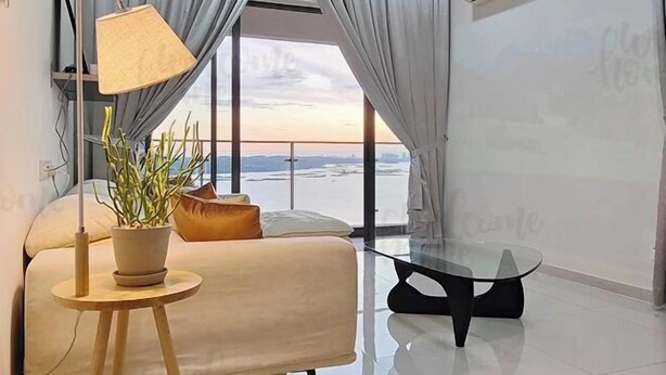 A modern living room with a sofa, a small round table with a lamp, and a view of the sea through large windows. - Amberside, Country Garden Danga Bay Homestay by Welcome Home (Johor Bahru)