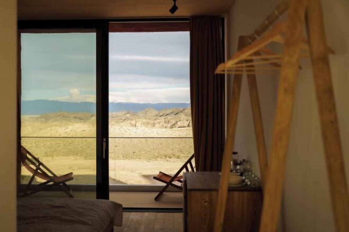 A room with a bed, a chair, a wooden desk, and a large window with a view of a desert landscape.