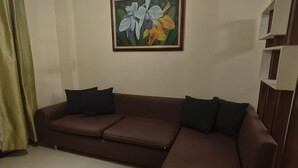 A brown sofa with black cushions, a painting of flowers on the wall, and a white shelf unit. - Sarasota Genlex Condominium (Pasay)