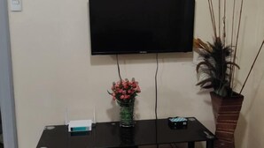A flat-screen TV mounted on the wall, a small table with a vase of flowers, and a tall potted plant beside the TV. - Sarasota Genlex Condominium (Pasay)