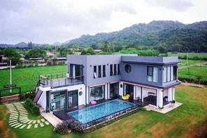 A modern two-story house with a swimming pool, large windows, and a view of a green landscape.