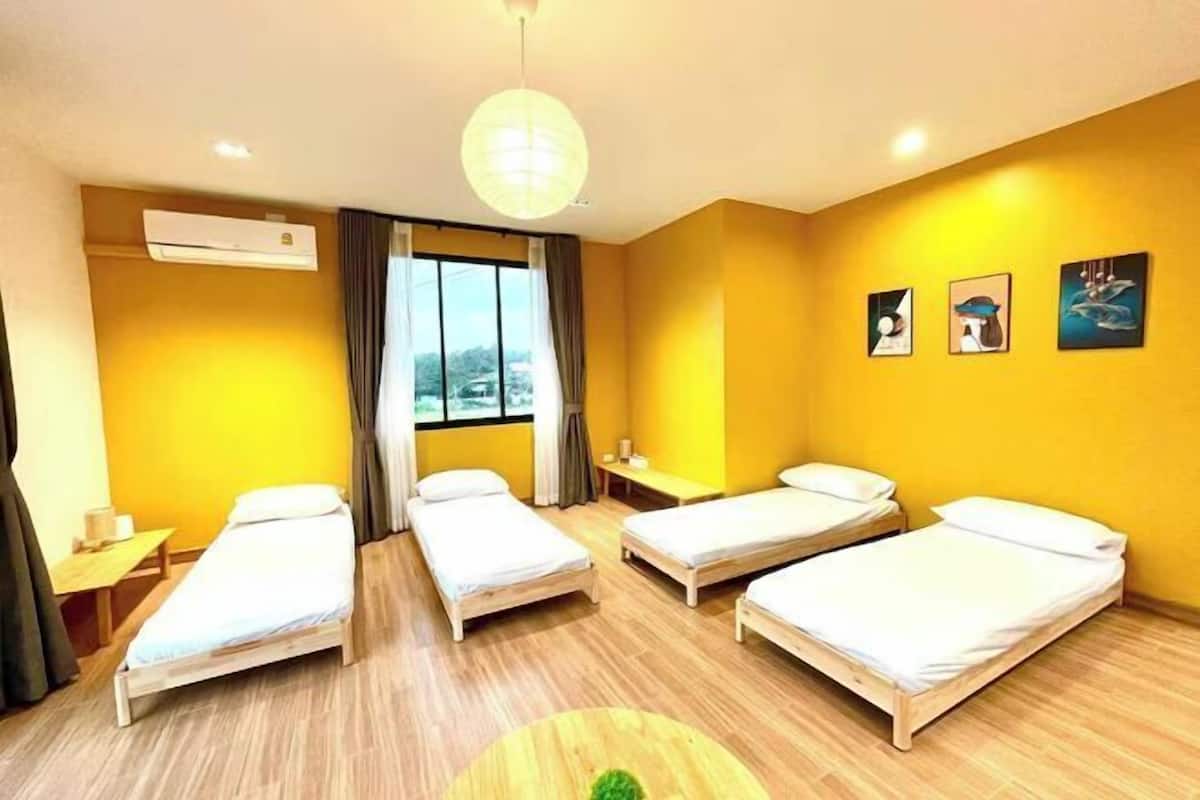 Une chambre d’hôtel avec quatre lits simples, un plancher en bois, une grande fenêtre et un ventilateur de plafond.