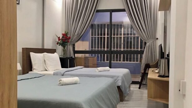 A hotel room with two beds, a small table with a vase of flowers, a desk with a computer, and a window with curtains.