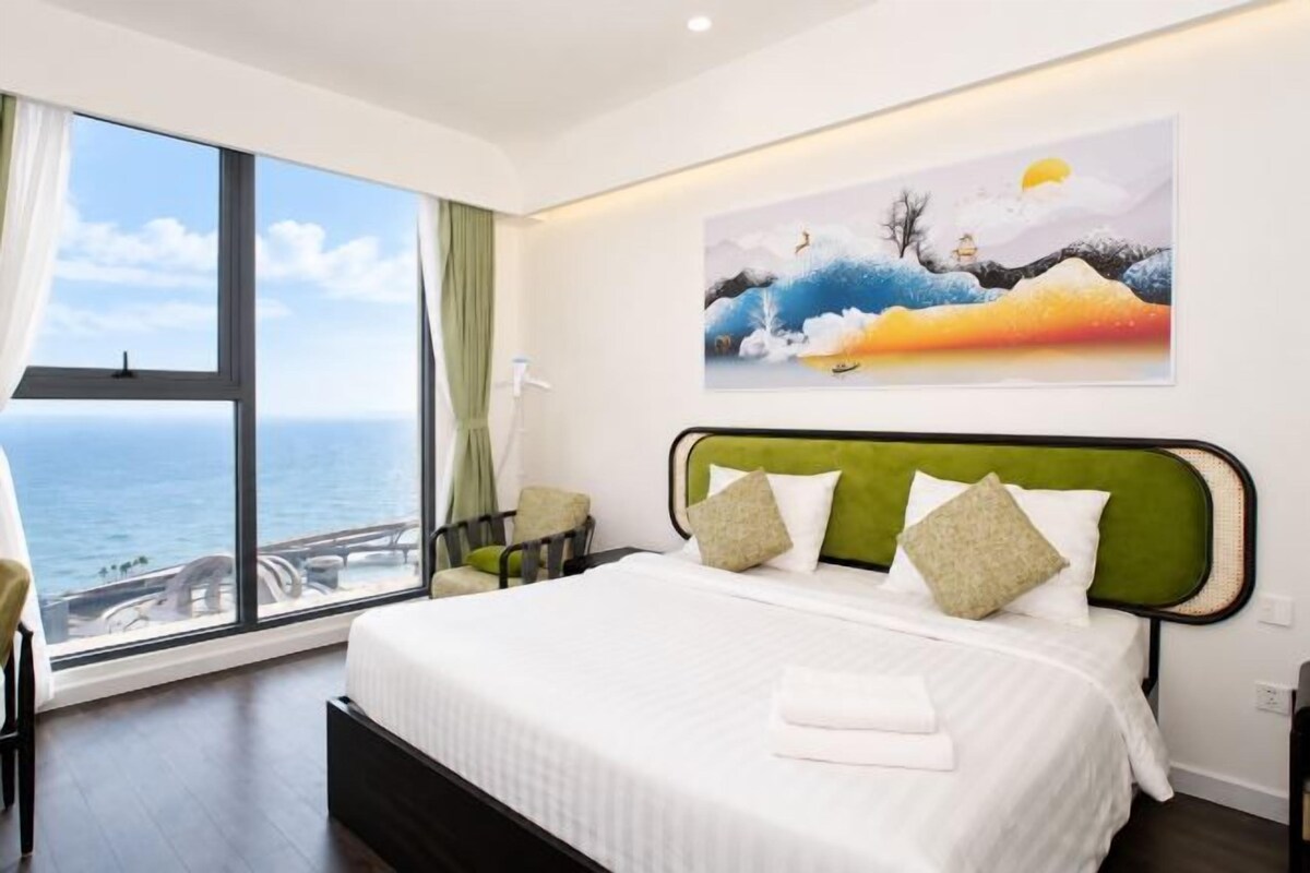 A hotel room with a large bed, a painting above the bed, and a view of the sea through a large window.