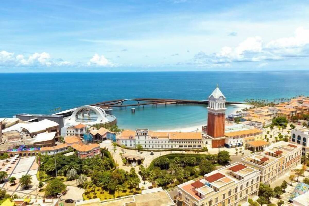 Un paisaje urbano costero con una torre destacada, una estructura circular grande y un puente sobre el agua.