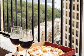 A window with a view of buildings and a pizza box with two glasses of red wine on a balcony. - Seaside Fireworks Apartment Free Pool & Gym (Phu Quoc Island)
