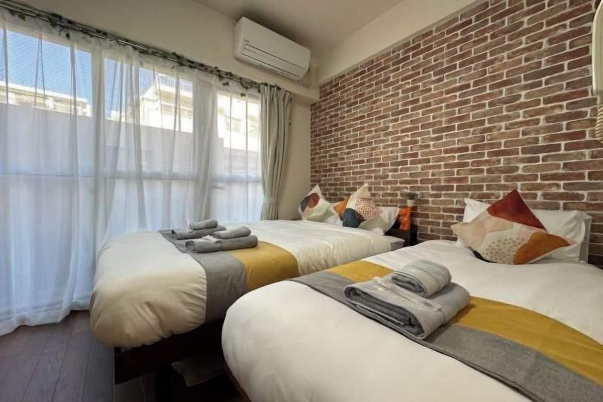 A hotel room with two beds, a brick wall, an air conditioning unit, and sheer curtains.