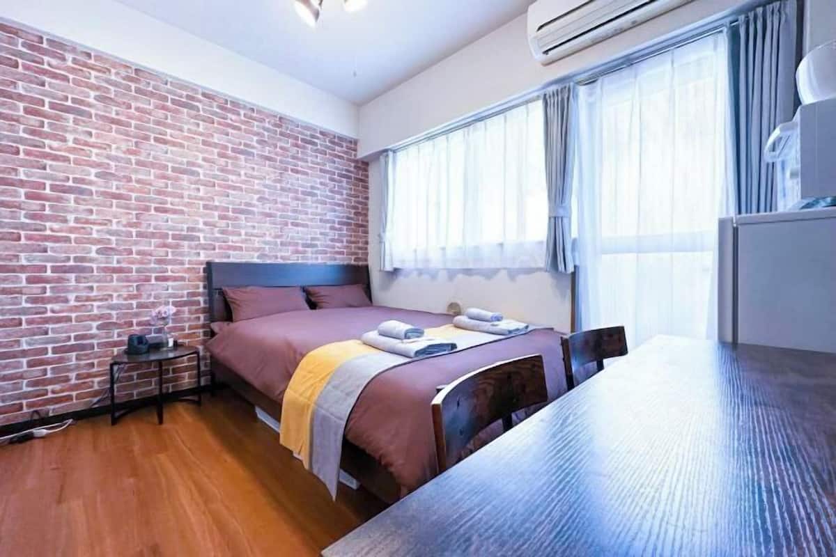 A room with a brick wall, a bed with a headboard, a bedside table, a window with curtains, and a wooden table.