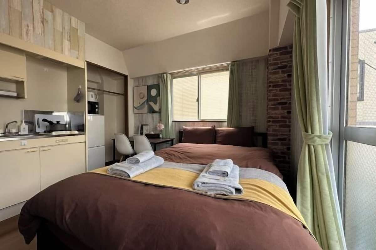 A hotel room with a bed, a desk, a chair, a window with curtains, and a kitchenette.
